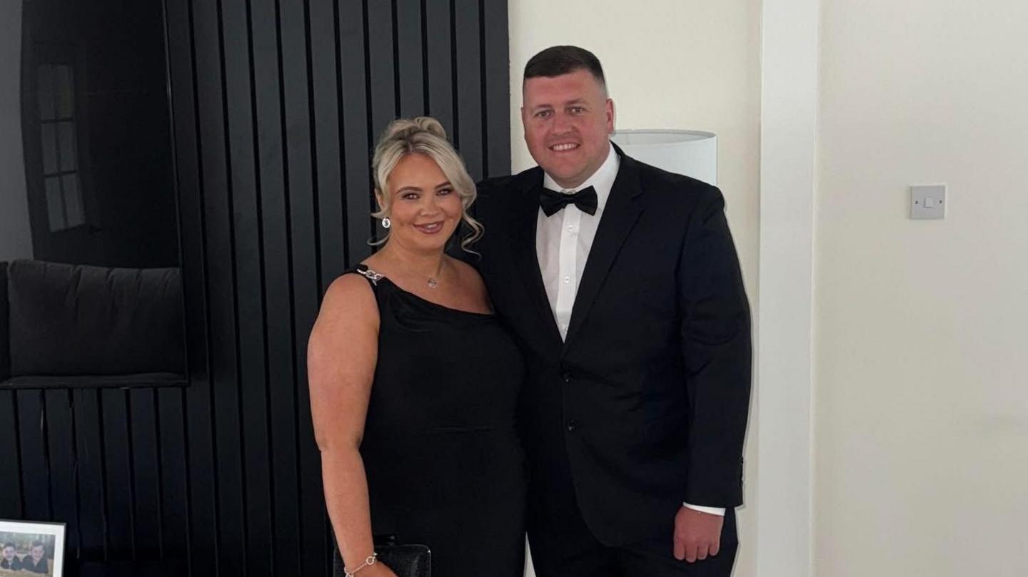 Chelsea McGillivray wearing a black dress with her partner Jack who is in a black suit, white shirt and black bow tie. They are standing together, smiling at the camera.