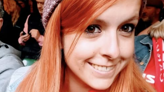Danielle is shown smiling and wearing a beanie hat in a crowd. Her hair is ginger coloured and she is looking at the camera with her brown eyes.