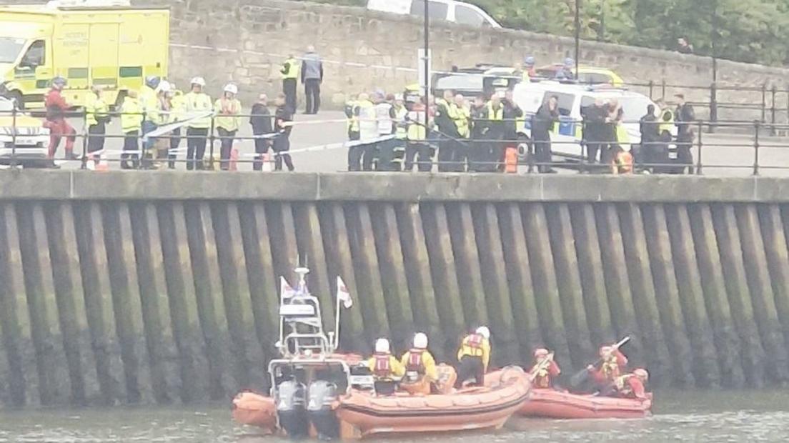 More than a dozen people, most wearing high-viz jackets are along the river bank in Sunderland, where a metal railing is missing and is covered in police tape There are emergency vehicles parked nearby and RNLI volunteers on two orange rescue boats in the river.