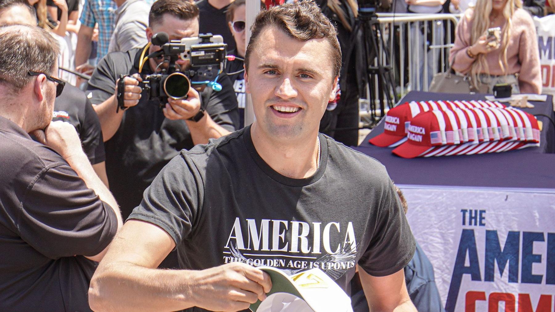 Charlie Kirk smiling at the camera wearig a T shirt with America written accross the front and crowds iand cameras n the background