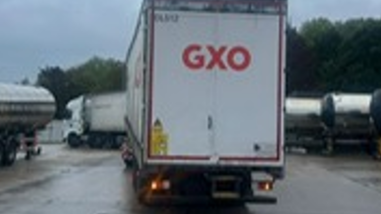 Rear view of white tri-axle curtain side trailers with branding for logistics company GXO on an industrial estate. Other HGVs are in the background.