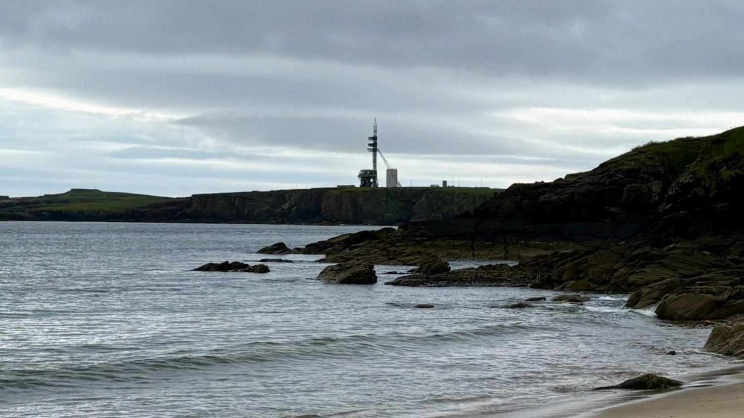 A different angle of the spaceport, seen this time from the beach. In the distance, a tall launch pad is on a long flat peninsula. There are waves and a small beach in front of the infrastructure.
