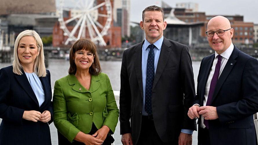 Four people are seen standing in Cardiff Bay - on the left a woman with blonde hair in a dark suit; a woman with brown hair in a green top; and two men in dark suits