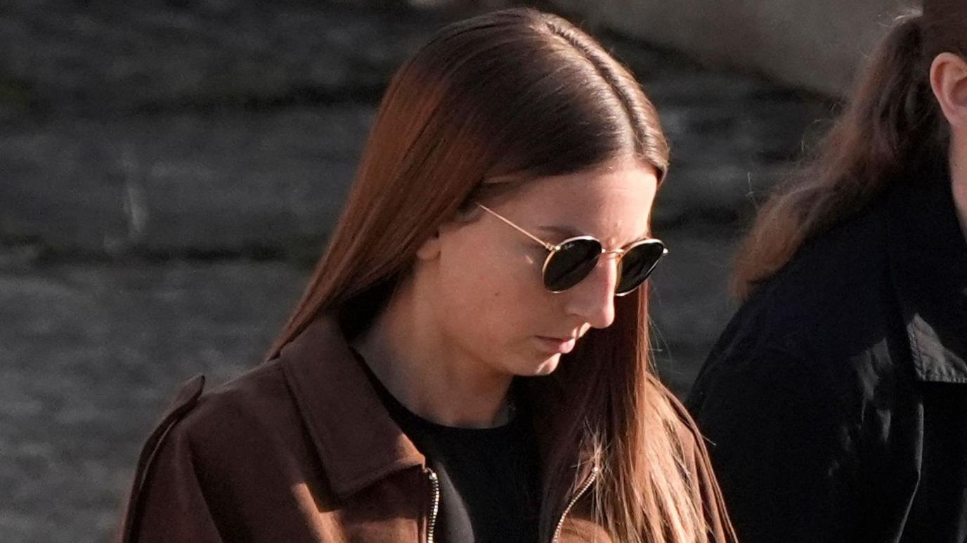Bronwen James walking into court wearing a brown jacket over black clothing and sunglasses with died long brown hair