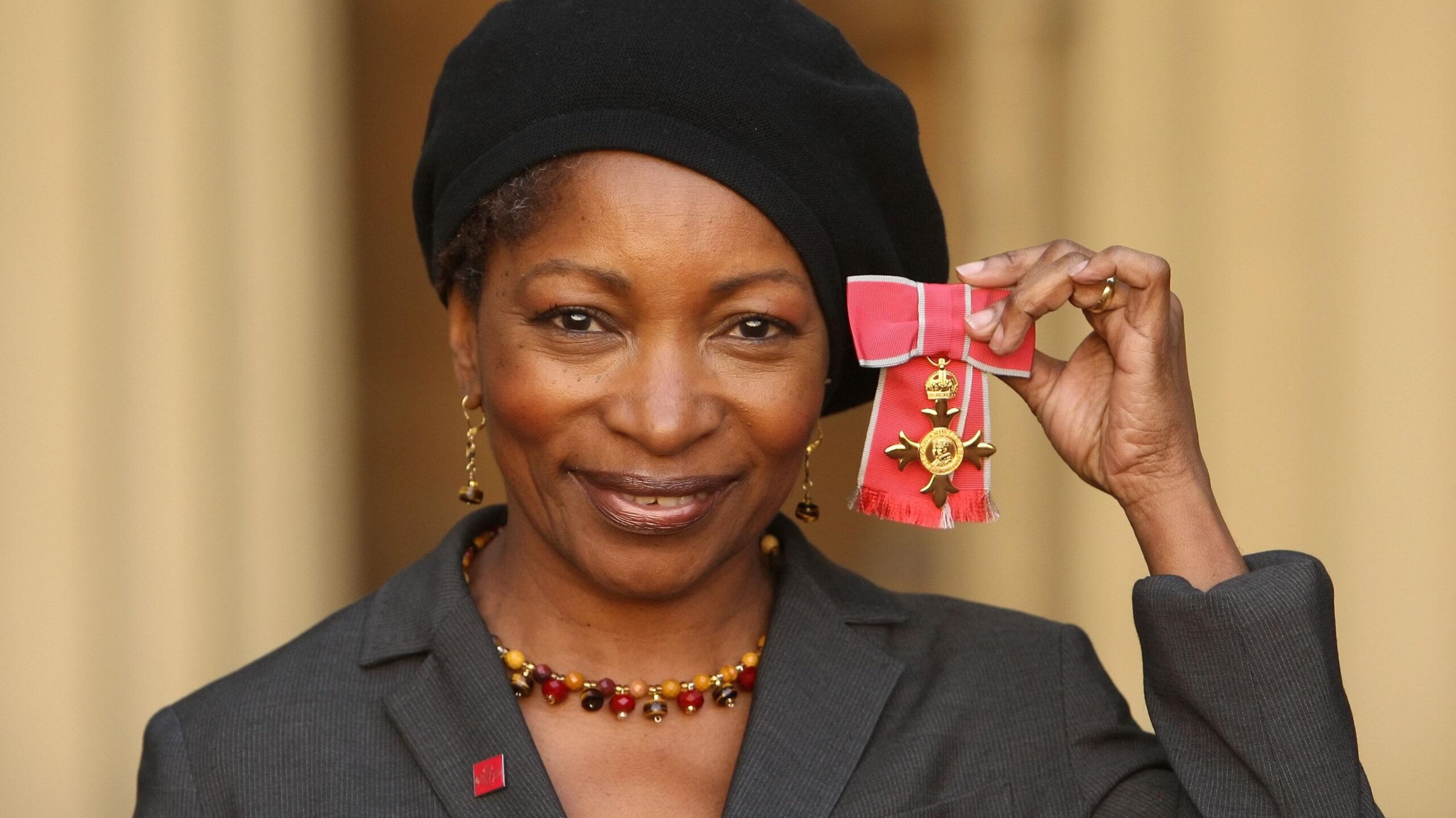 Bonnie Greer in smart grey suit, hat and necklace, smiling and holding up her OBE medal