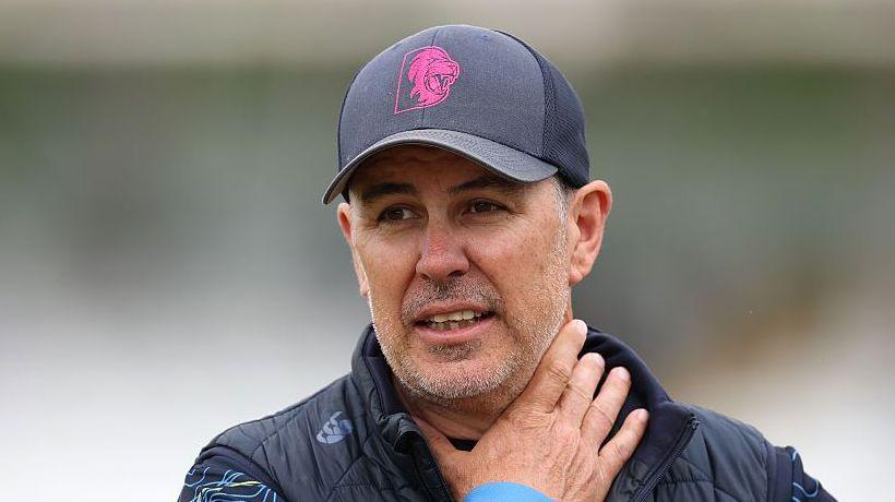 Ryan Campbell wearing a Durham gilet and navy blue cap with his right arm clutched up towards his neck