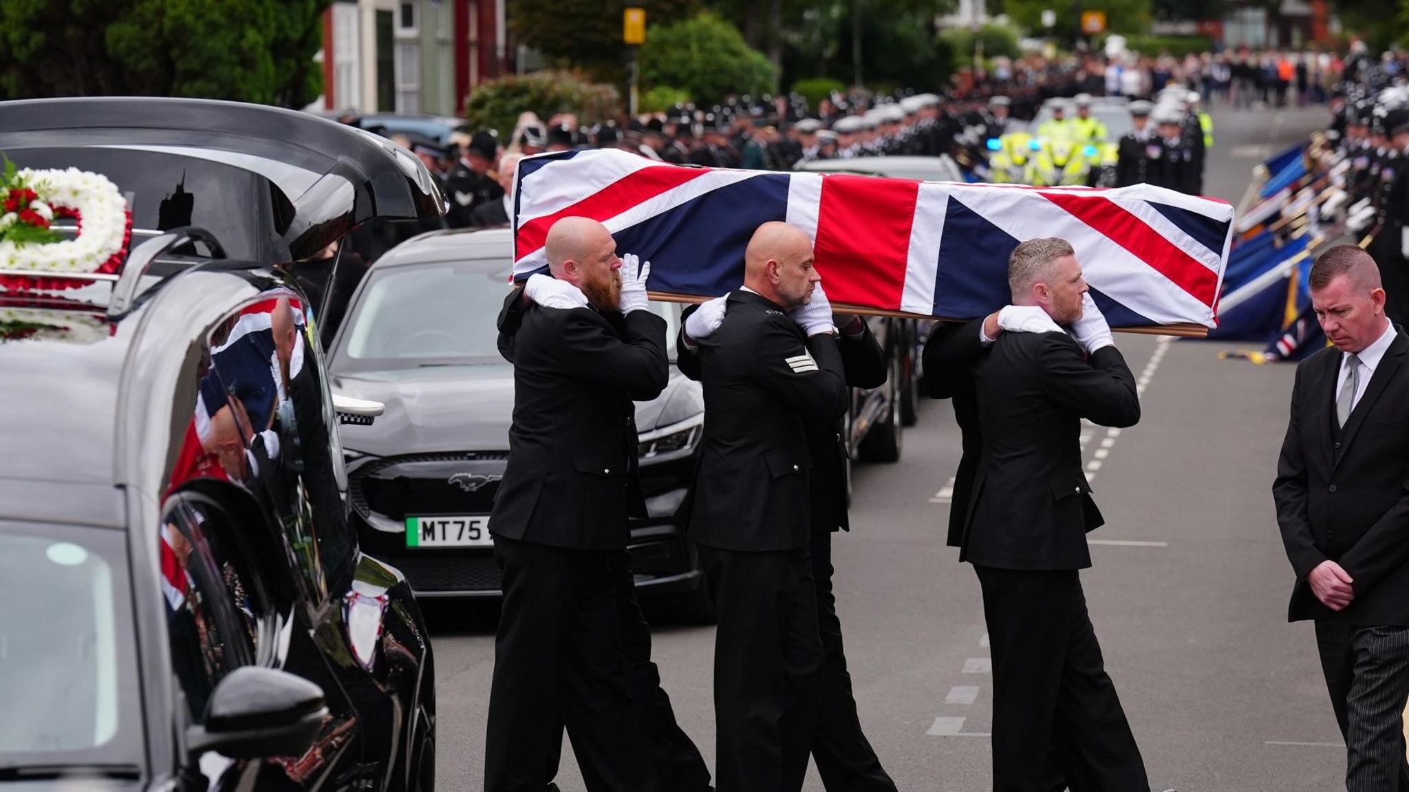 Six police officers in black suits and white gloves carry PC Blades' coffin on their shoulders. It is draped in a union flag. Only three of the men's faces are visible and they all have sombre looks. The hearse is to the left with the boot open and other black cars are parked behind it. In the distance there are police on motorbikes in yellow hi-vis with crowds lining the street, also out of focus. There is a man in a black suit jacket, grey trousers and silver tie with his hands clasped together on the right, in front of the coffin. He has brown, shaved hair and is looking down.