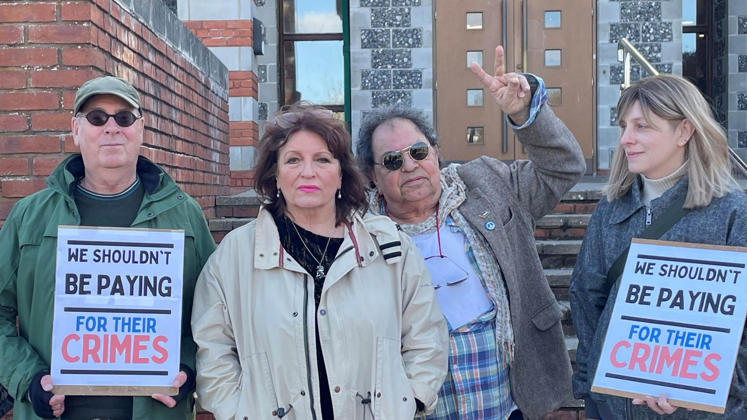 Four people stood in front of a court. Two of them are holding signs reading - "We shouldn't be paying for their crimes".