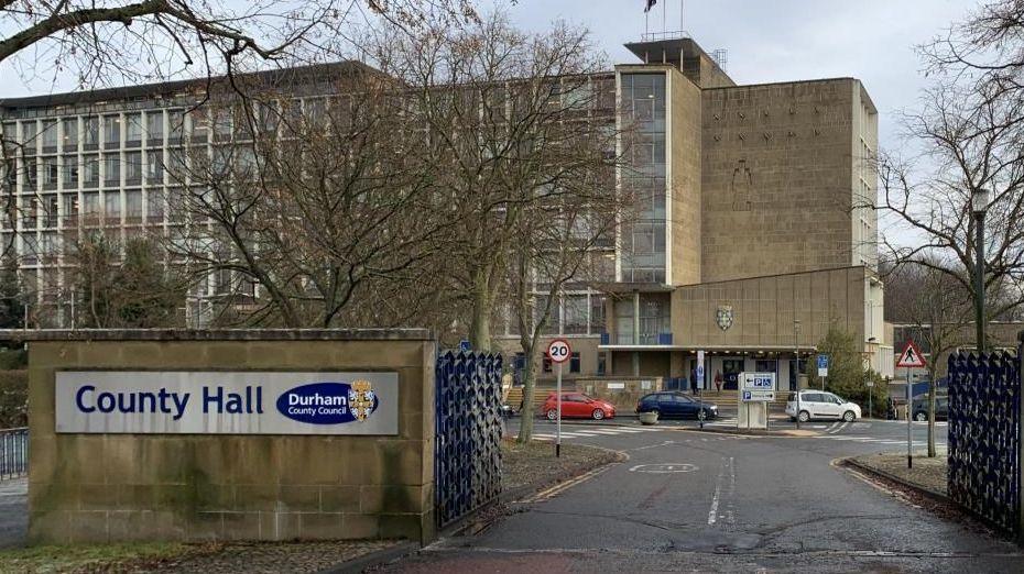 Durham County Hall is a multi-storey beige stone building. Its facade is mostly covered in windows. A blue gate leads into the building. Several cars are parked in front of its entrance. A grey and blue sign with the local authority's crest and reading County Hall is placed on a stone fence next to the gate.