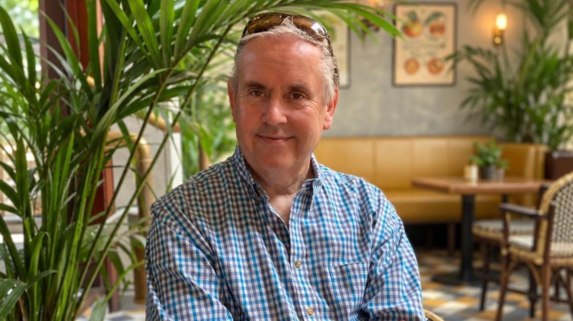 Dave Talbot smiles at the camera as he sits in a restaurant. He has brown sunglasses resting on top of his head and wears a blue checked shirt. There is an indoor plant to his left.