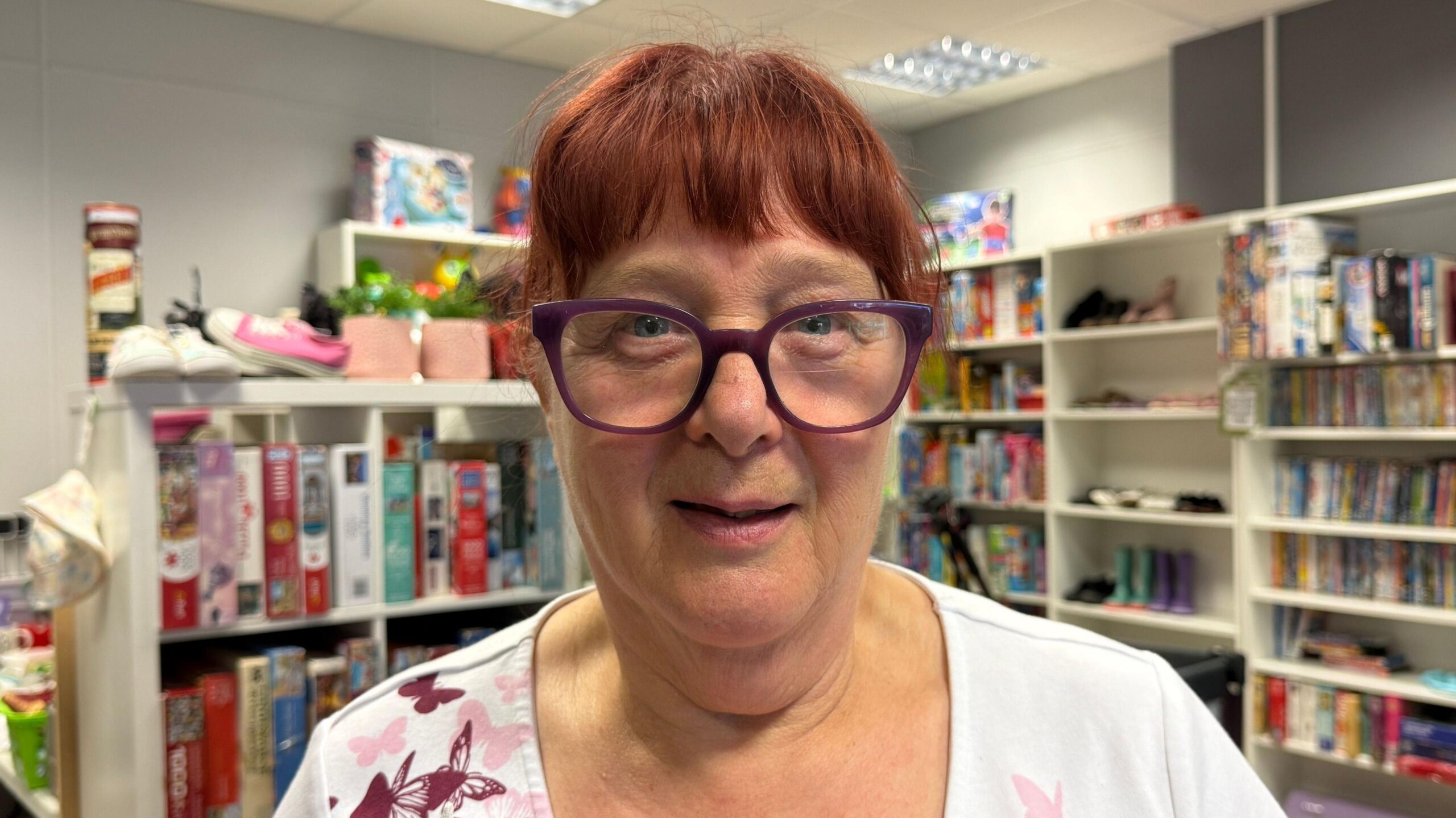 A woman is standing in front of bookshelves in a shop. She is wearing a white top with pink butterflies on it. She has big purple glasses, blue eyes and red hair.