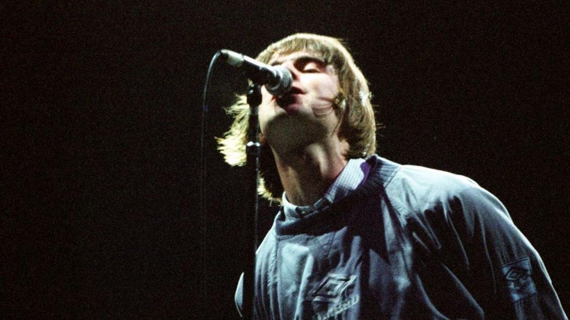 Liam Gallagher wearing an Umbro jumper while singing into a mic during one of Oasis's famous Maine Road gigs in 1996