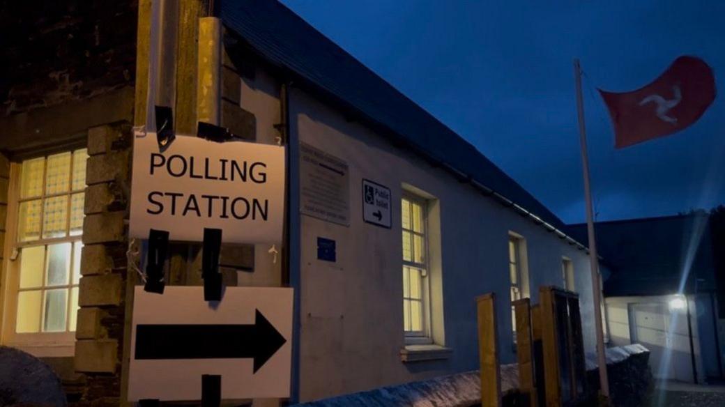 A sign reads polling station with an arrow, there is a Manx flag flying in the background.