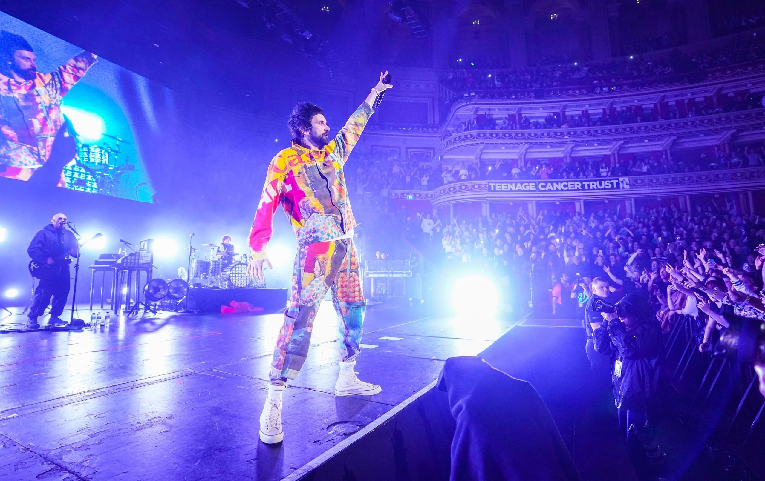 Kasabian on stage during the Teenage Cancer Trust charity shows of 2023. Serge is wearing a multicoloured jumpsuit, raising his left hand towards the sky. In the front row, dozens of fans are reaching their arms towards the stage