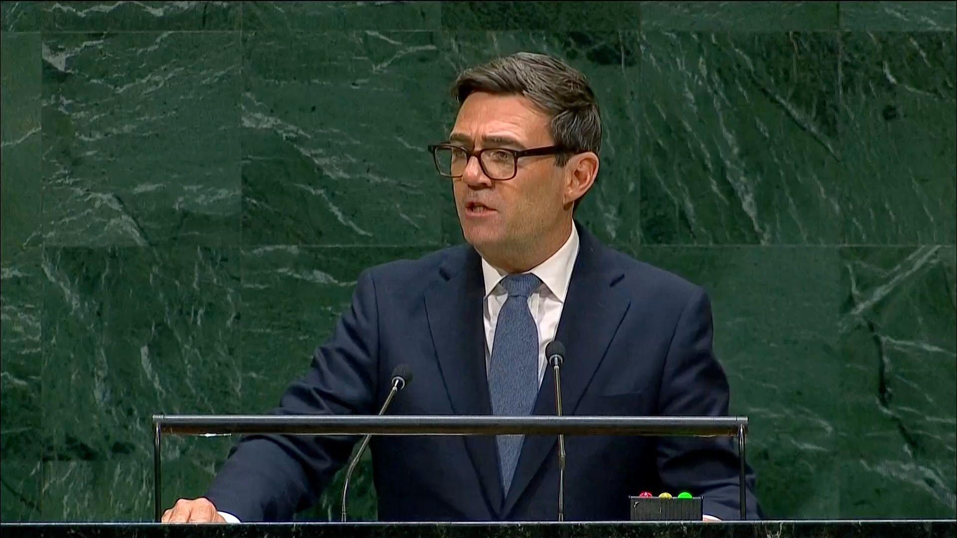 The British Prime Minister Andy Burnham speaking at a lectern in a navy suit in front of a green tiled wall.