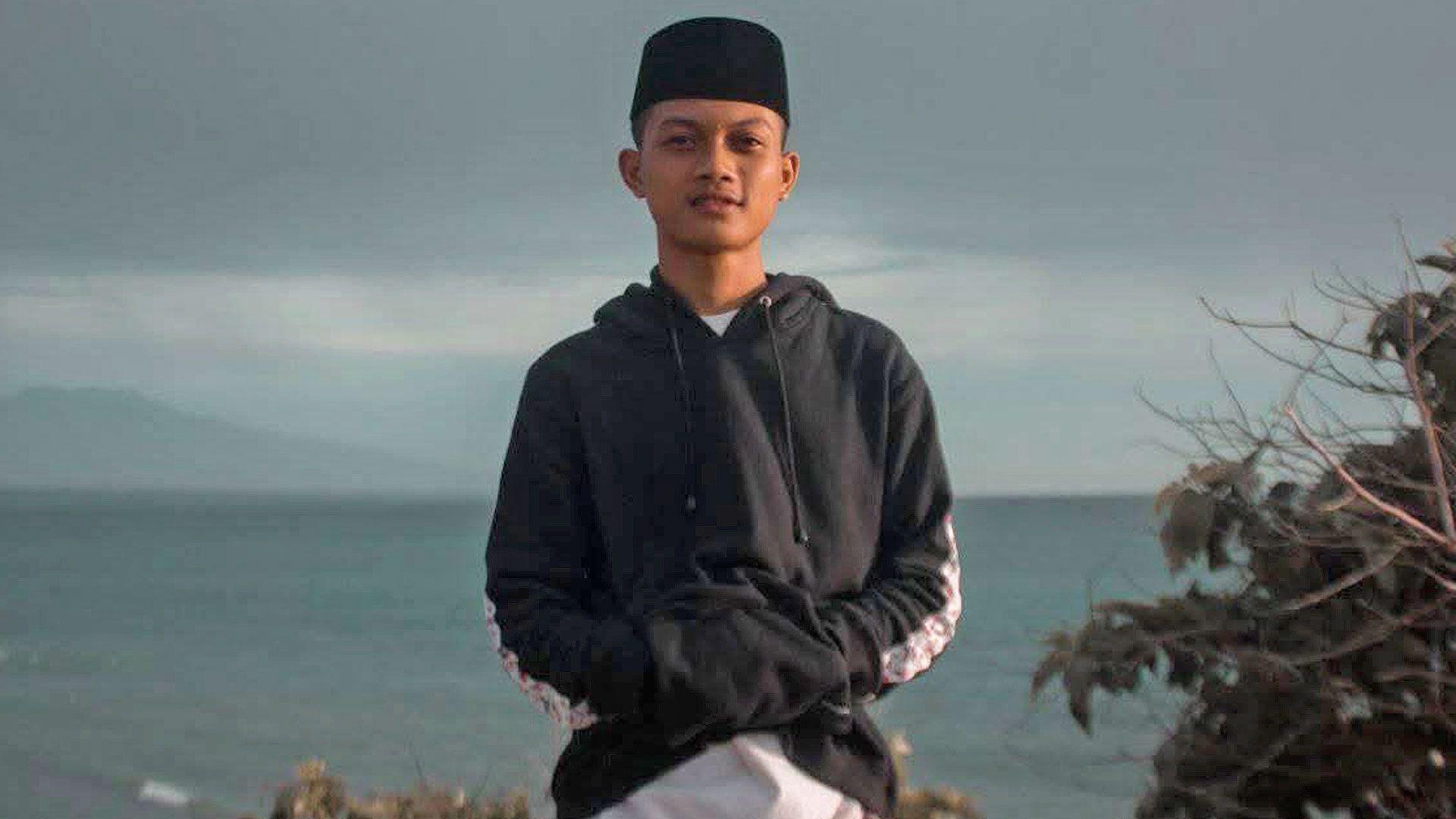 Yusron stands looking at the camera with the sea as a backdrop. He is wearing a black hoodie and white trousers, and a black kopiah on his head. He has his hands in his pockets.