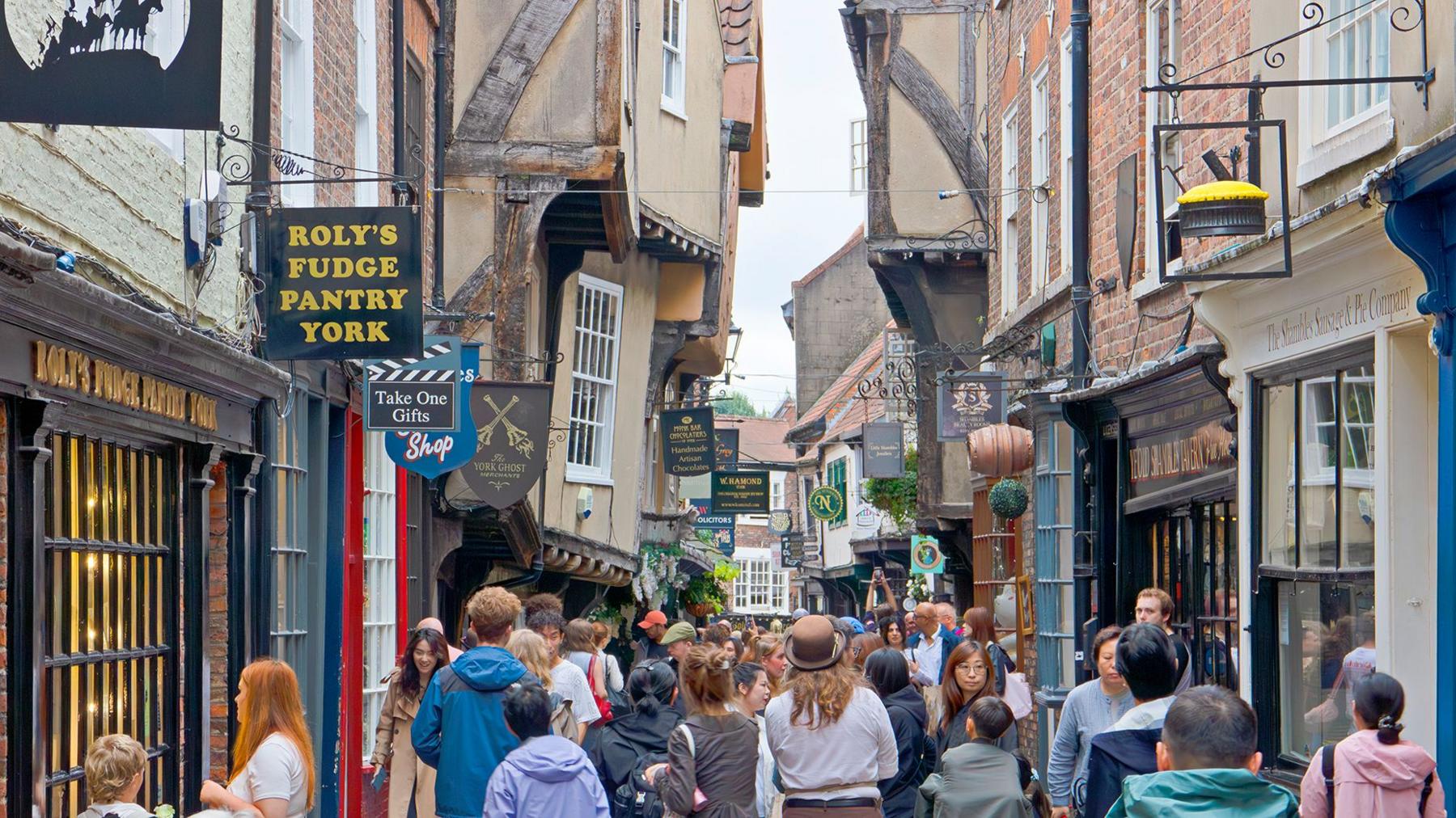 View along The Shambles in York, a narrow medieval street with timber-framed buildings housing shops and cafés. The scene shows the busy pedestrian area that retains its characteristic overhanging upper stories and historic layout.