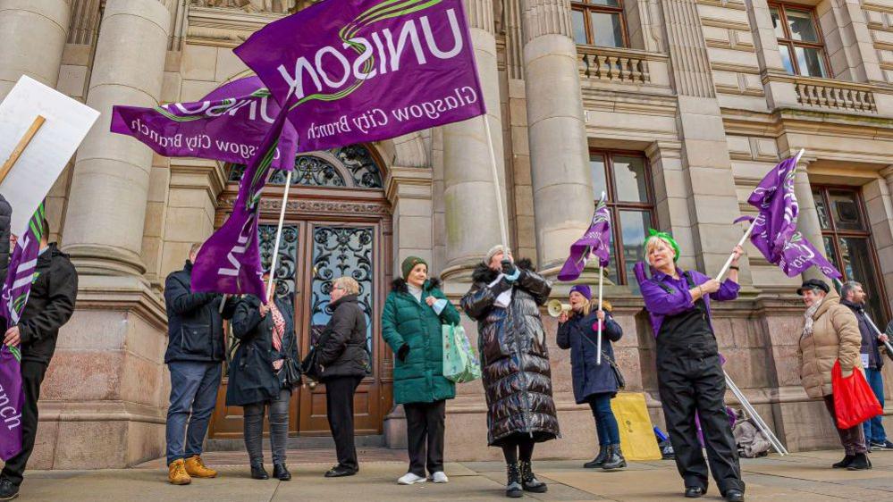 a group of workers protesting and waving purple 'Unison' flags outside an Glasgow City chambers.