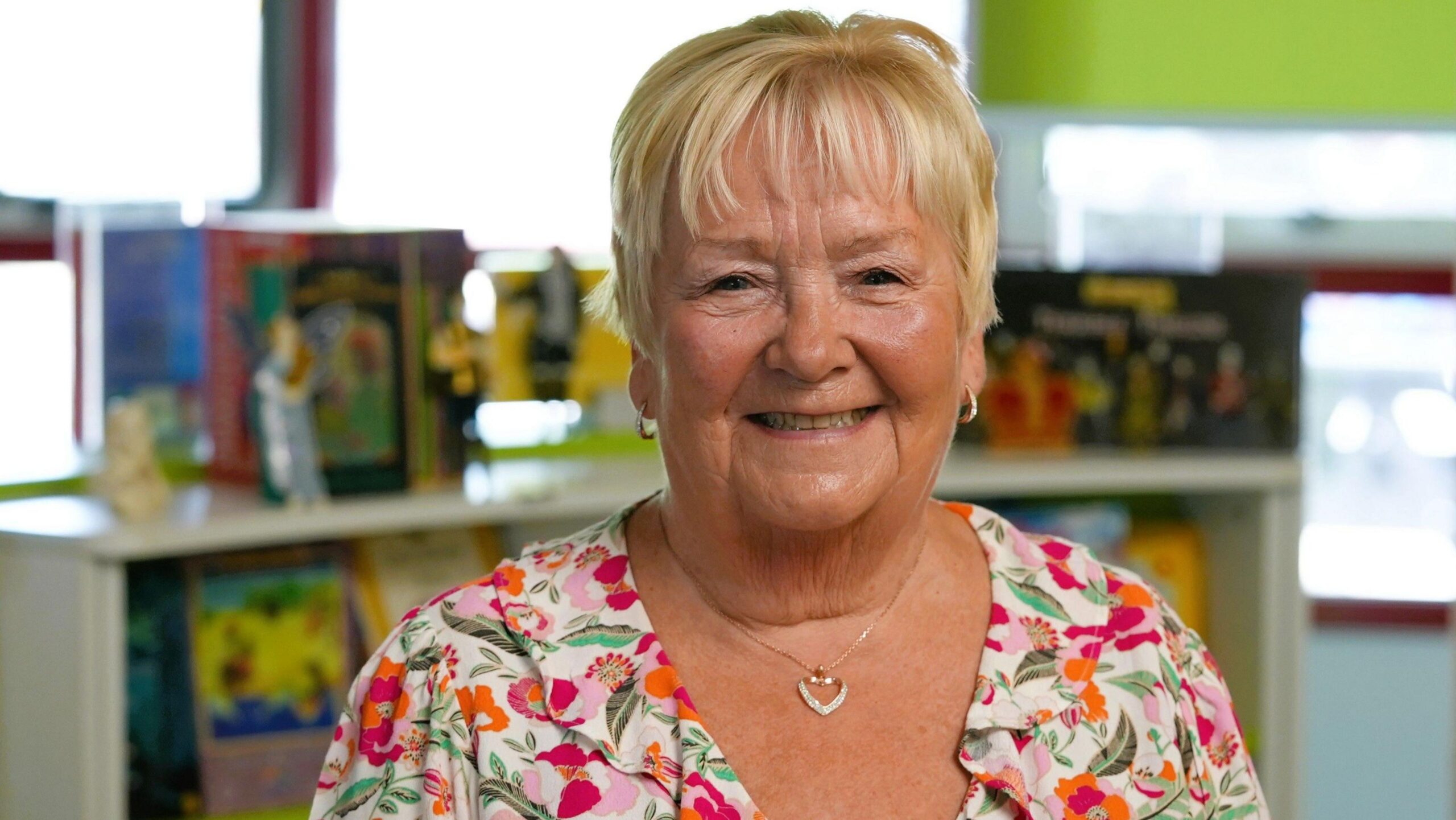 Denise, wearing a floral dress and with short blonde hair, smiles at the camera. It is a head and shoulders shot.
