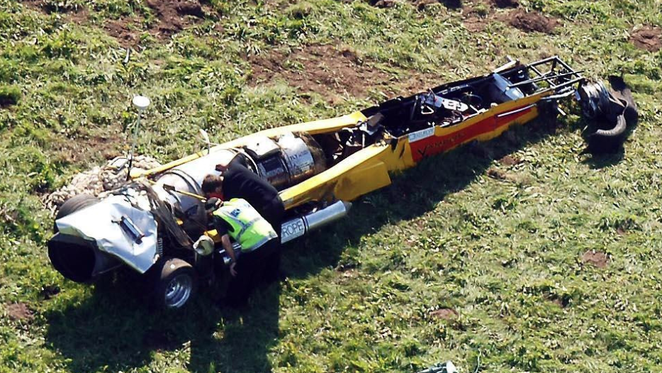 A long red and yellow dragster-style car stands stationary with major damage to its front end, including missing wheels. Two men, one wearing a police jacket, examine the jet engine at the rear of the vehicle.
