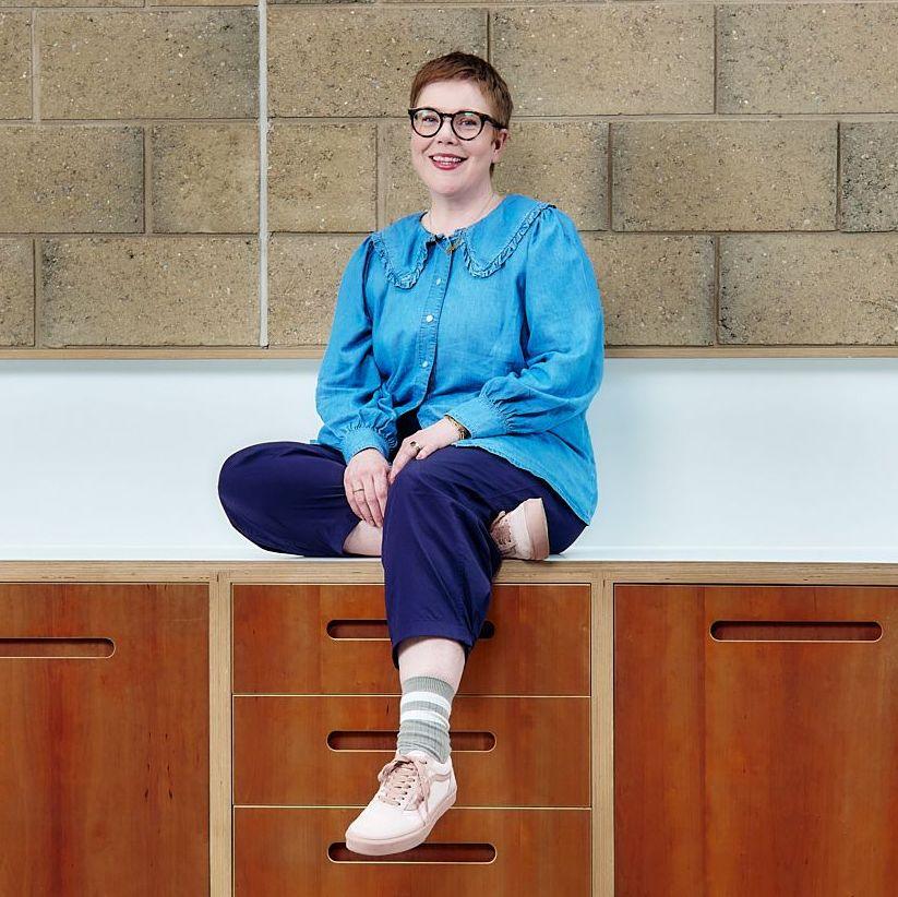 Anna Moynihan, wearing a blue shirt, blue trousers, stripey socks and trainers, sits on a kitchen worktop. She is smiling at the camera and is wearing glasses.