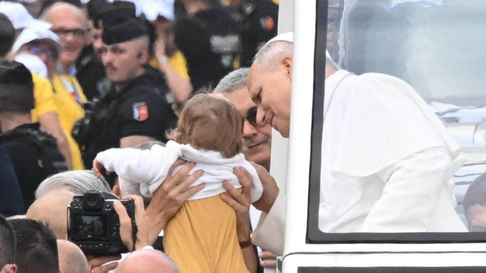 The pope leans over to look at a child being held up to him. He is in his popemobile and crowds can be seen around them. A number of police officers can bee seen nearby.