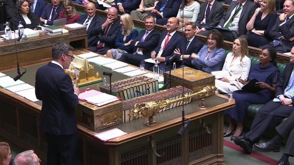 Andy Burnham is stood at the despatch box in the House of Commons, with a red folder containing pieces of paper in front of him. His back is to the camera and he is facing Conservative MPs sat on the green benches opposite. Burnham is wearing a dark coloured suit, white shirt and glasses.