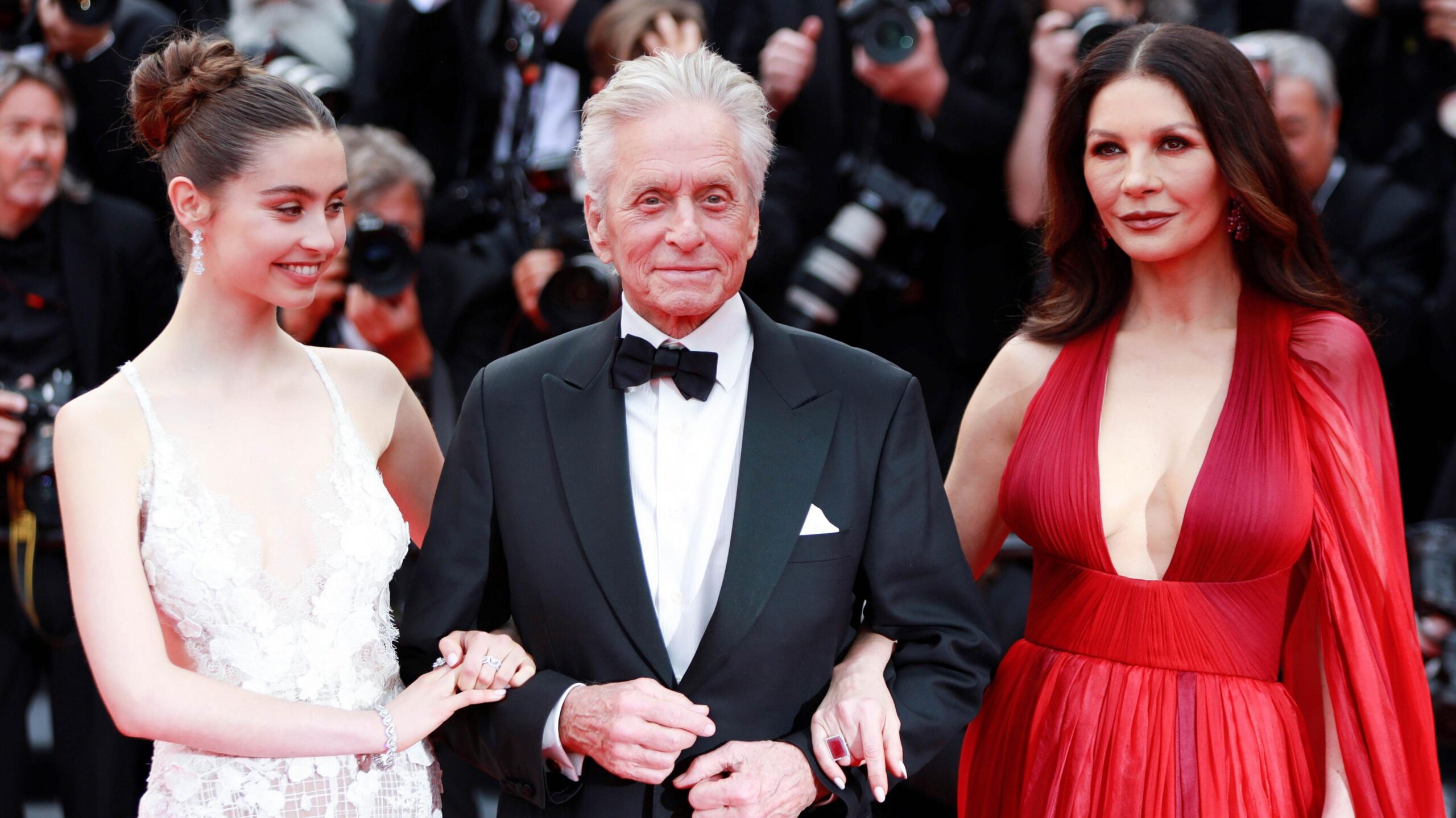 Carys Zeta Douglas, Michael Douglas and Catherine Zeta-Jones are photographed on a red carpet together. Carys is wearing a white lace dress, with her hair up. She is linking arms with her father, Michael Douglas, who is wearing a black suit and white shirt. On his other arm is his wife, Catherine Zeta-Jones, who is wearing a red dress and her hair down.