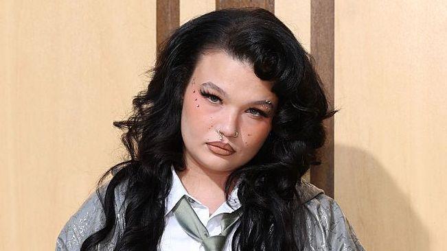 Lola Young at a fashion event. The singer wears her long dark hair loose and tilts her head towards the camera. She is styled in a grey jacket over an unbuttoned white shirt and green tied.
