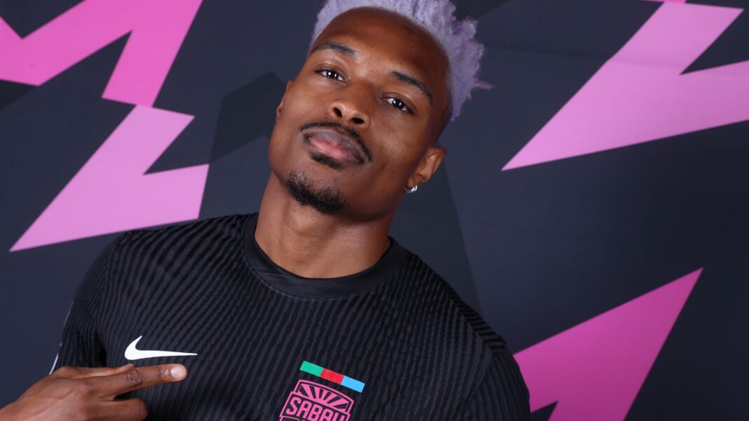 The Rwandan eyeing Champions League history against Man Utd Joy-Lance Mickels, with hair dyed a light pink colour and styled extravagantly by being pushed high above his head but shaved entirely at the sides, is visible from chest up wearing a black Sabah shirt. He points with his right index finger to the bright pink club badge which reads 'Sabah' on his left breast as he looks into the camera. He stands against a black background bearing pink zigzag patterns reminiscent of lightning bolts