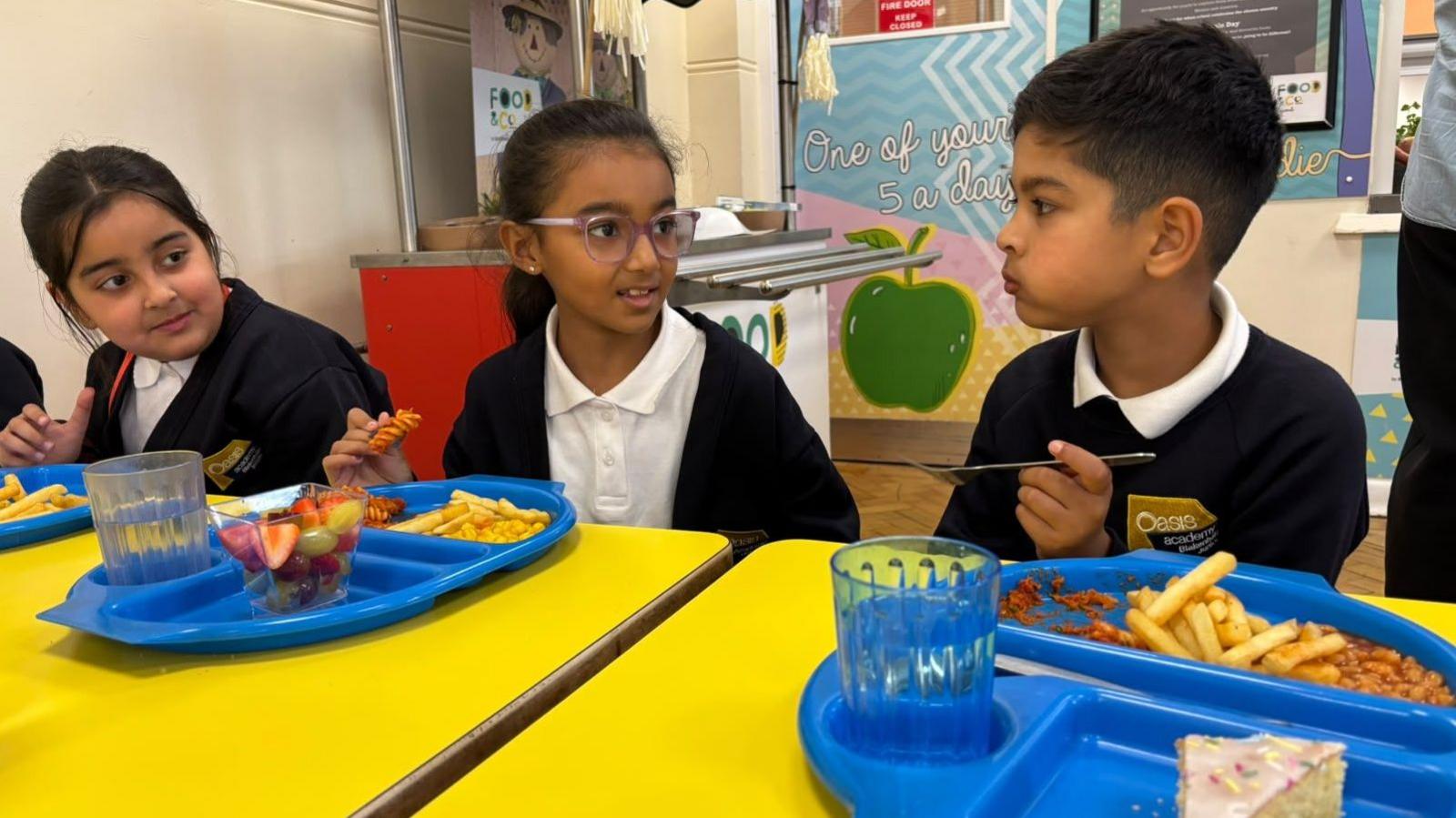 Three primary school pupils in uniform sit at bright yellow tables in a school canteen, eating lunch from blue trays with chips, fruit and dessert while talking together.