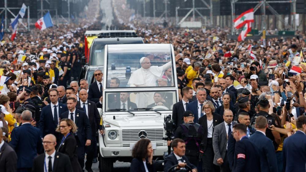 Pope Leo waves from his popemobile as he moves down the Champs-Élysées Avenue ahead of the open-air mass. Thick crowds can be seen on both sides of the road as some people hold up their children to be blessed while others hold up their mobile phones. Large flags can be seen being waved in the background.