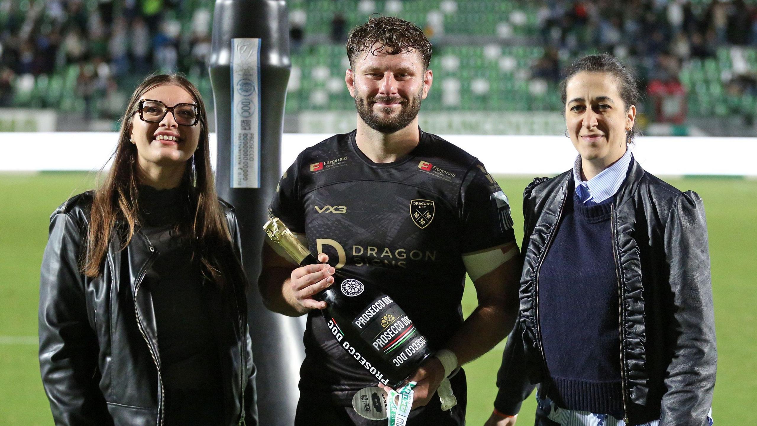 Thomas Young is presented with a giant bottle of prosecco after winning man of the match for Dragons at Benetton