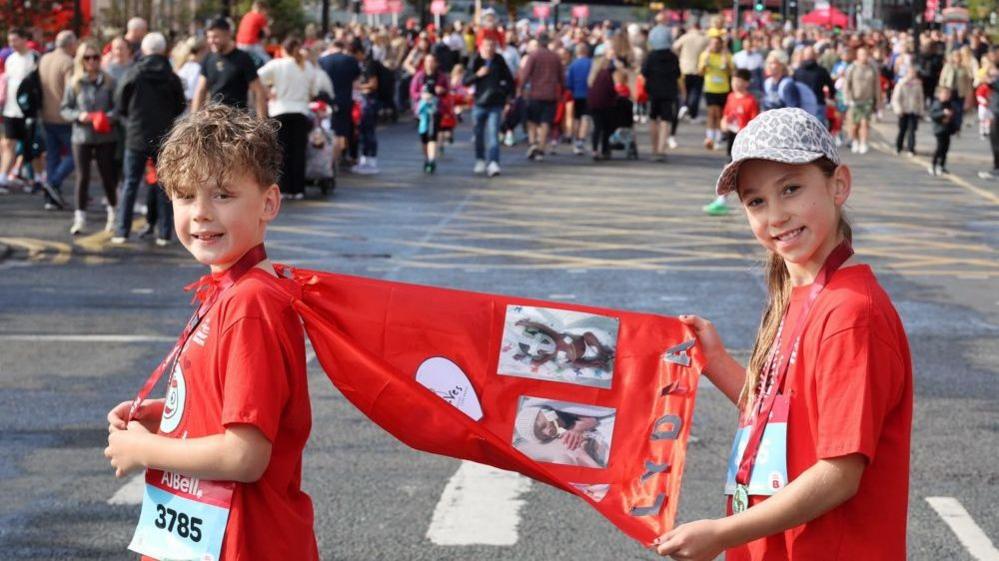 Two youngsters are wearing red running gear. One of them is wearing a red cape with pictures stuck to it and letters which spell out "Lydia". The other child is holding the cape up. They are both smiling at the camera while crowds gather on the grey tarmac road behind them.