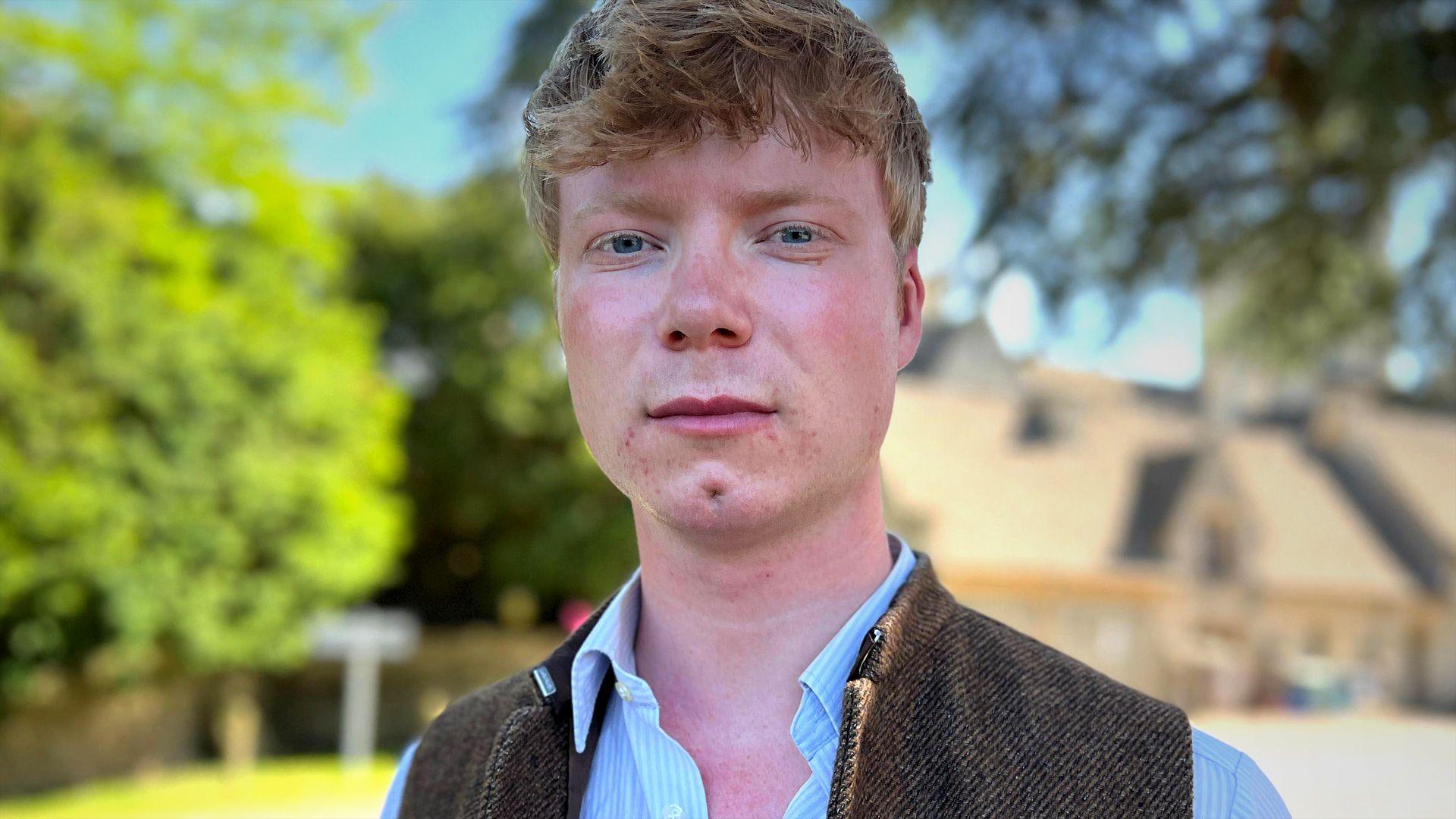Lewis Jackson is pictured looking directly at the camera with a serious expression. He is a young man with short strawberry blonde hair and is wearing a white and light blue striped shirt with a tweed gilet over the top. He is surrounded by trees on a sunny day.