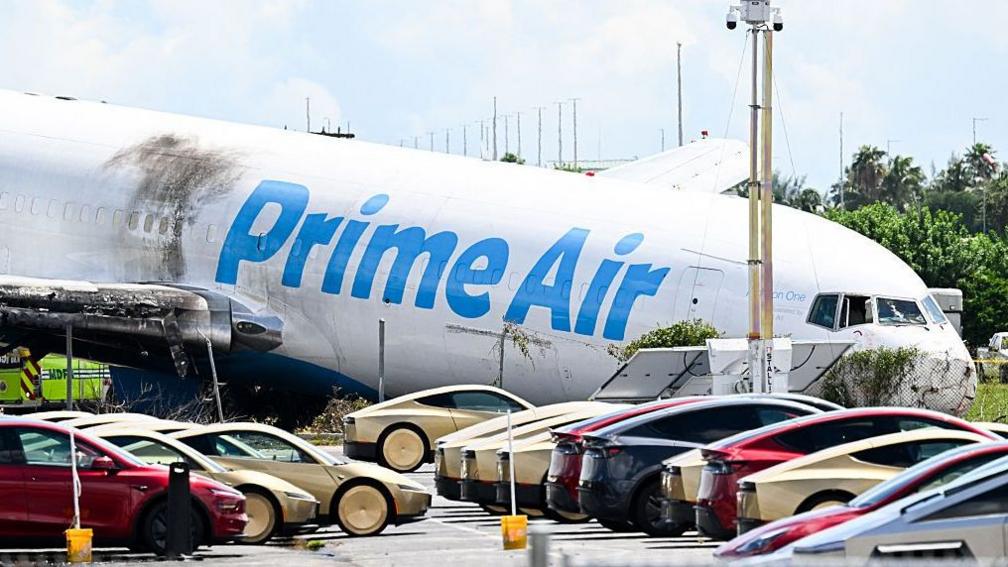 A parking lot filled with Tesla Cybercabs is seen in the foreground as the nose of a Prime Air cargo plane rests on the ground after the aircraft overran the runway and struck cars the previous day at Miami International Airport in Miami, Florida, on September 7, 2026.