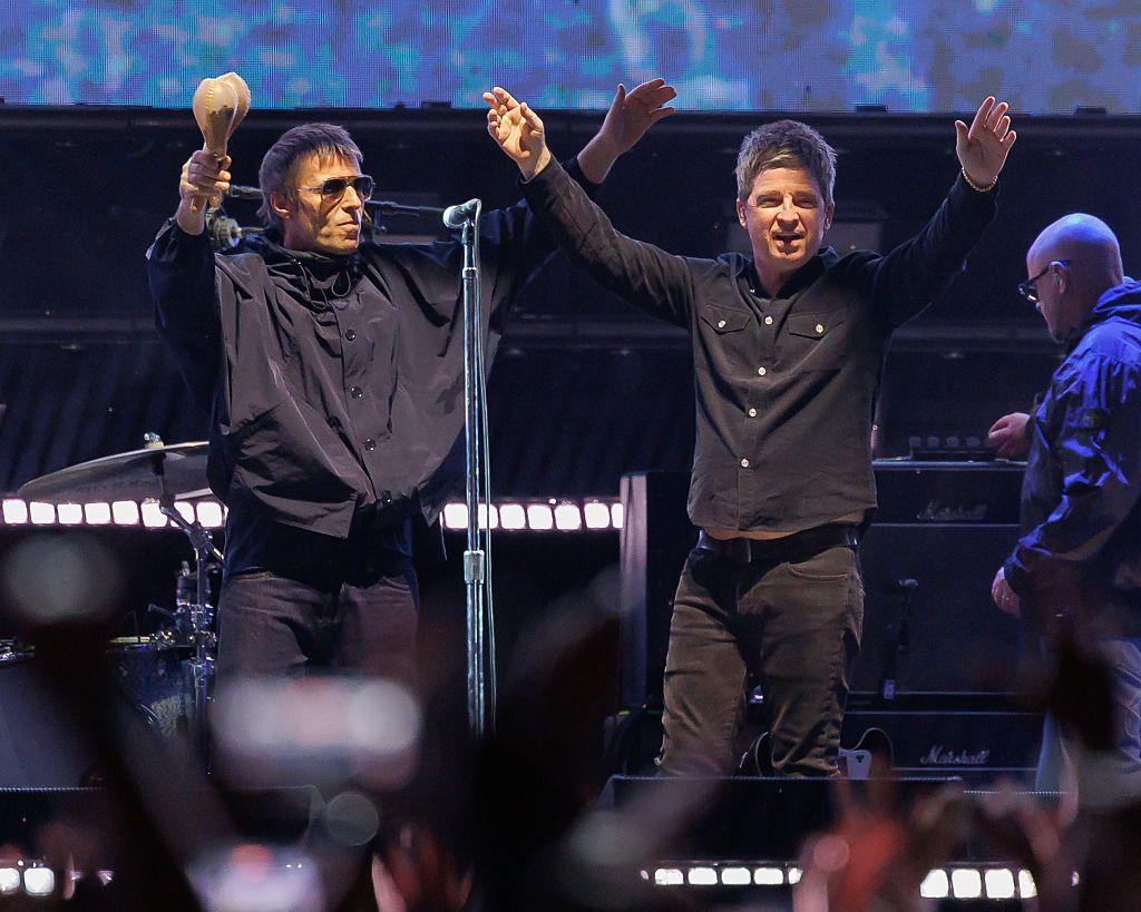 Oasis on stage in 2025. Both Liam and Noel are wearing black shirts and raising their arms into the air to greet fans. Noel is holding a pair of maracas in his right hand.