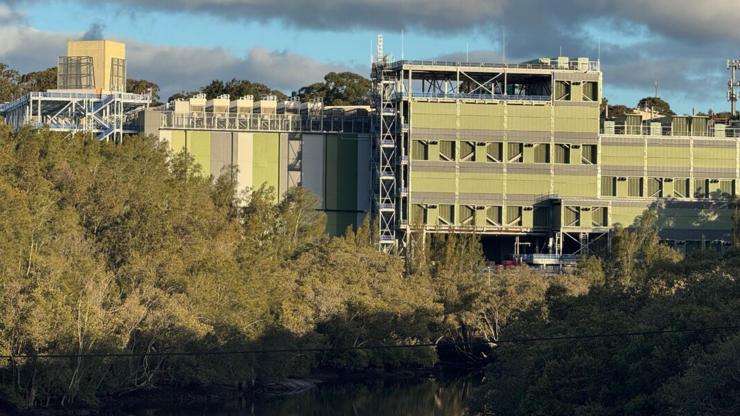 Data centres are booming in Australia – but at what cost? A large green and grey building situated amid bushland and a river down below.