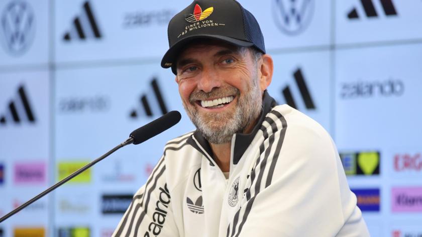 Germany boss Jurgen Klopp, wearing a white jacket and black baseball cap, smiles during a news conference with a microphone in front of him