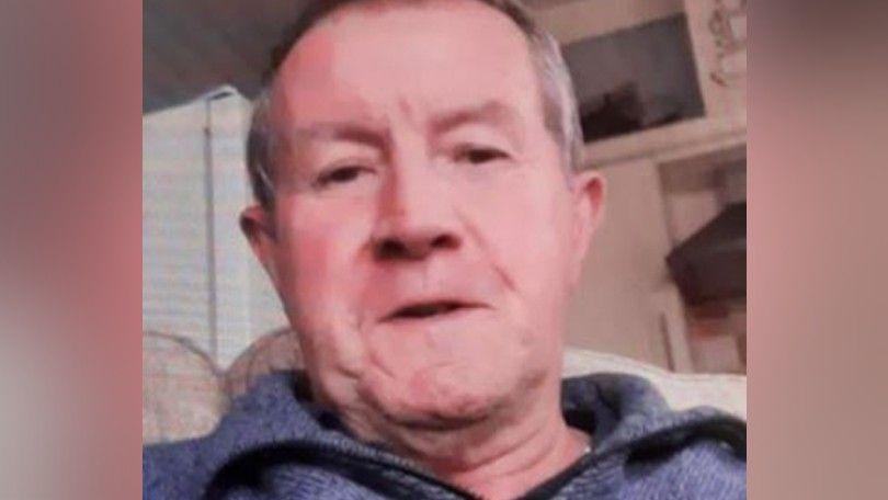 A low-quality image of Richard Scatchard. He is an older man with short grey hair and is wearing a grey top with wide lapels