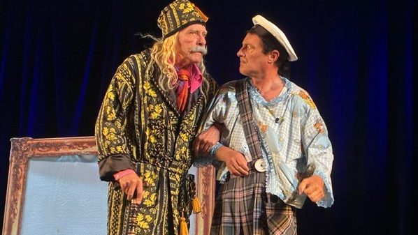 Two men act in a theatre - one is wearing a dressing gown on the left, the other wears a colourful blue and white shirt