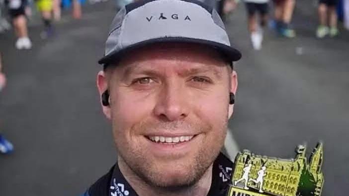 PC Blades takes a selfie with a medal for completing a run in Middlesbrough. The medal shows the gothic Town Hall building with the outline of two runners in front of the building. He has neat stubble and is smiling, with a grey running cap on his head. He has black earphone buds in his ears.