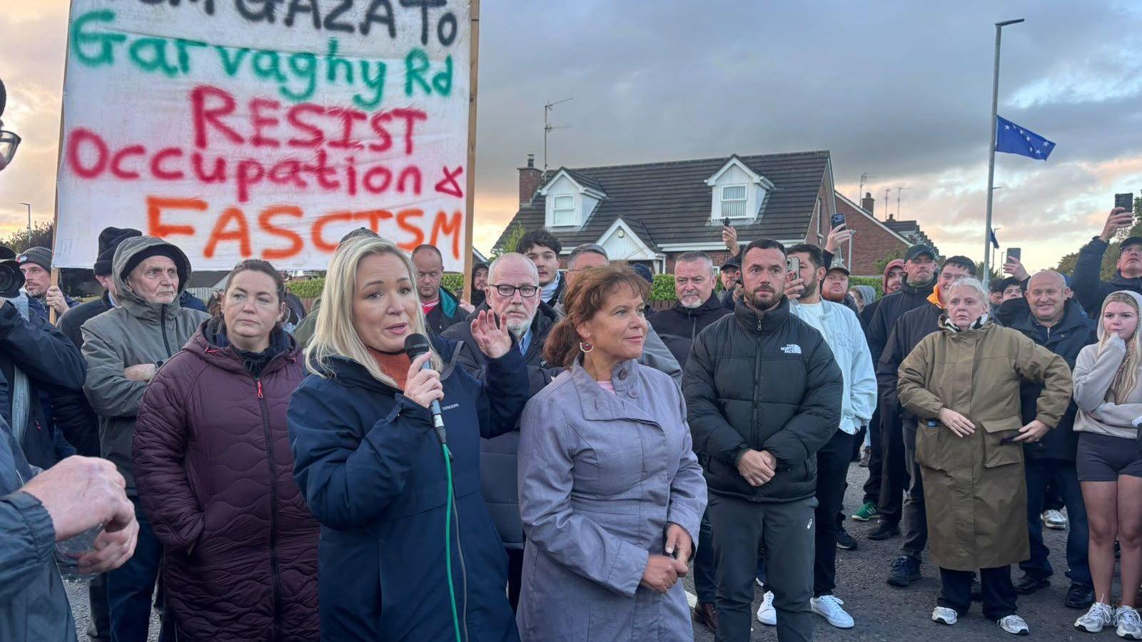 A blonde woman speaking on a microphone, she is joined by numerous protesters, there is a banner calling for Garvaghy Road to resist occupation