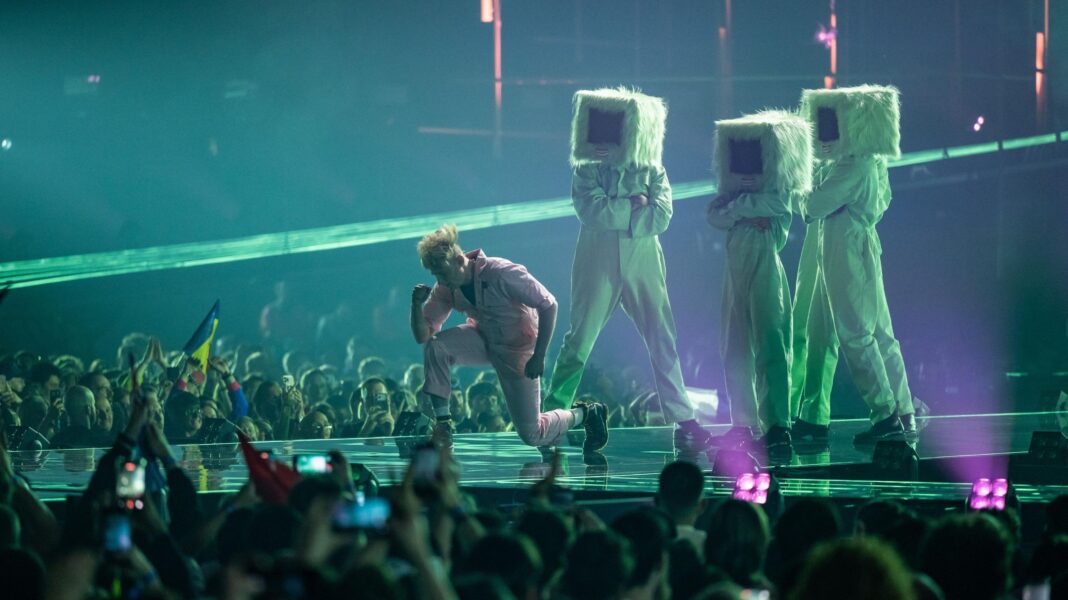 A performer in a pale pink outfit kneels on a Eurovision stage holding a microphone while singing. Behind are three large pale green furry characters with rectangular black screens instead of faces. The characters wear matching pale green outfits and stand in a line behind the performer. Purple and green stage lighting fills the arena, while audience members wave flags and hold up mobile phones around the edge of the stage.