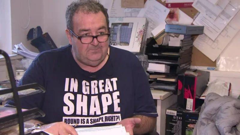 Gerard Woodhouse looks at the camera while wearing glasses and a t shirt saying 'in great shape, round is a shape right?' while sitting in an office