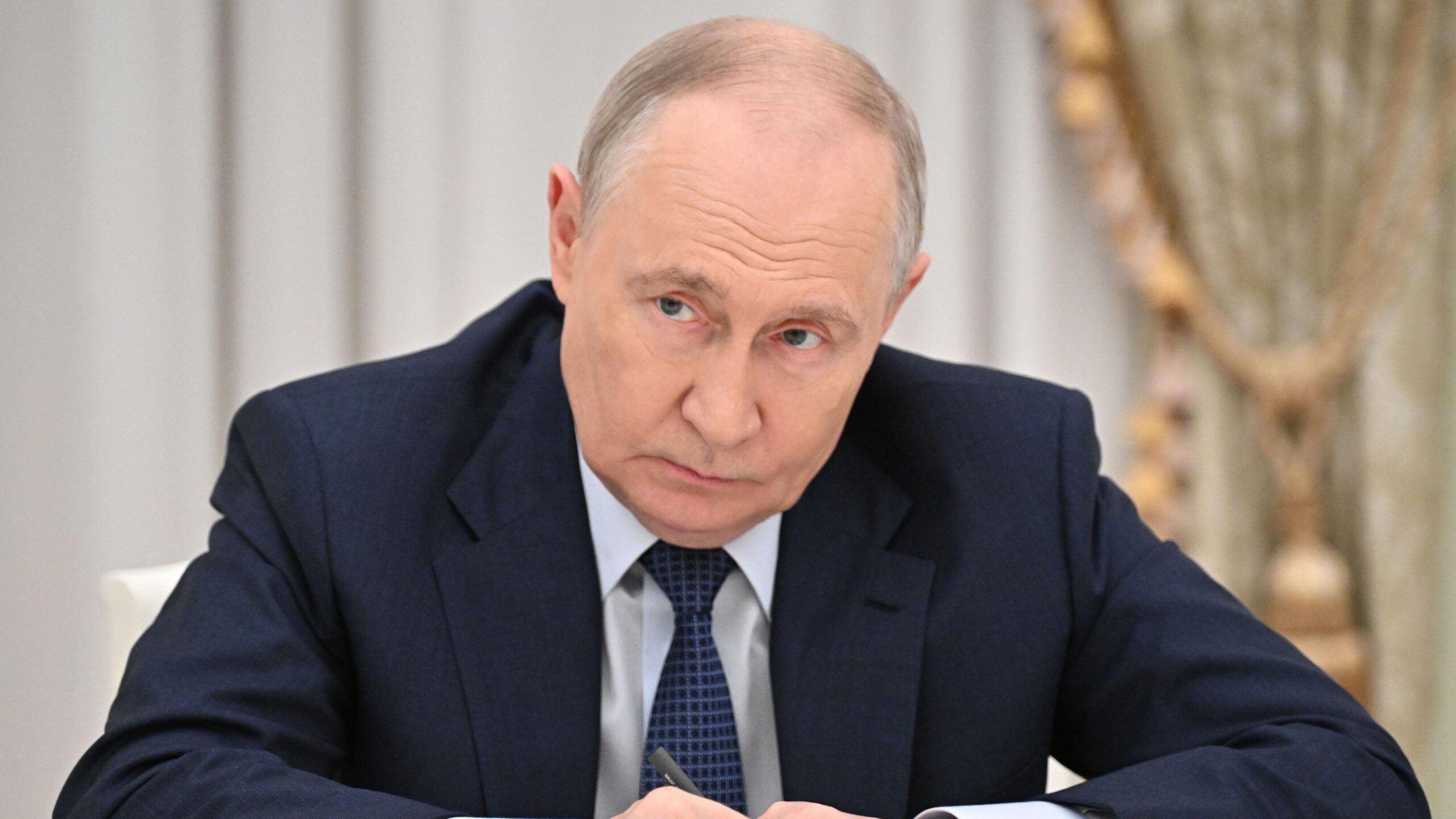 Vladimir Putin holding a pen with his arms outstretched, wearing a blue suit, blue tie and white shirt