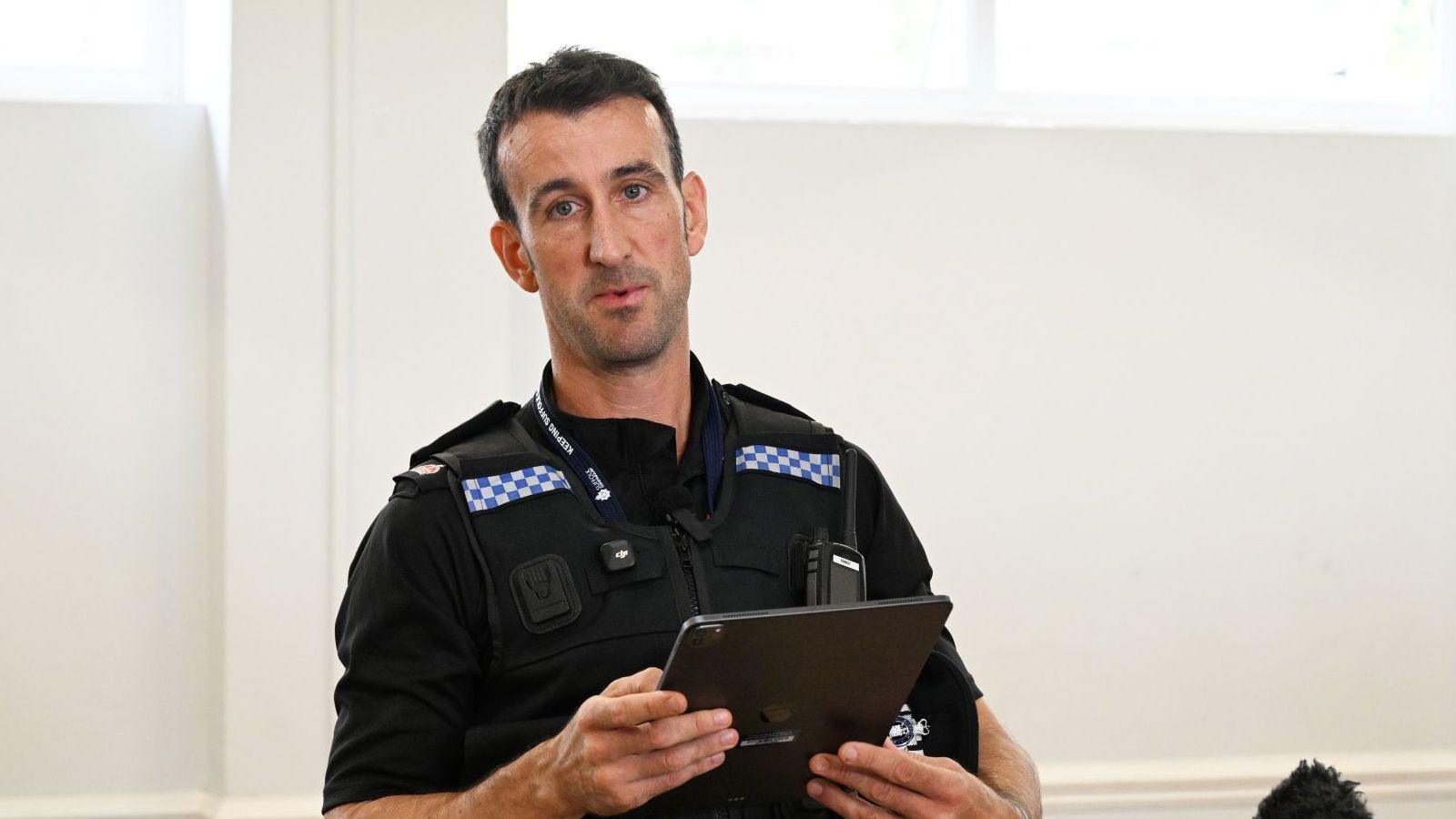 A police officer reading out a statement while holding an iPad or tablet with a black cover on it. He is wearing his police uniform and has black hair