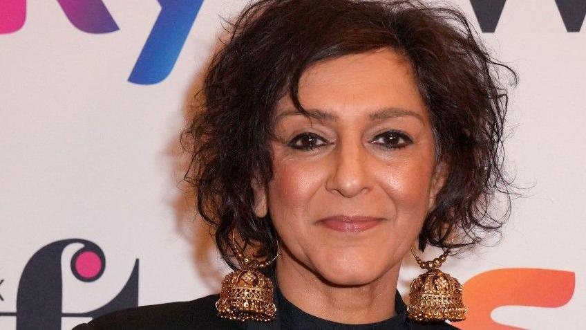 Meera Syal is looking at the camera, in front of a wall with brand names on it. She has her hair up and is wearing large gold earrings,
