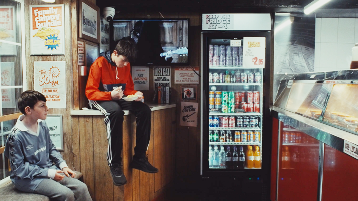 Still from a video showing two boys sitting in a chip shop with a fridge full of drinks and a number of posters