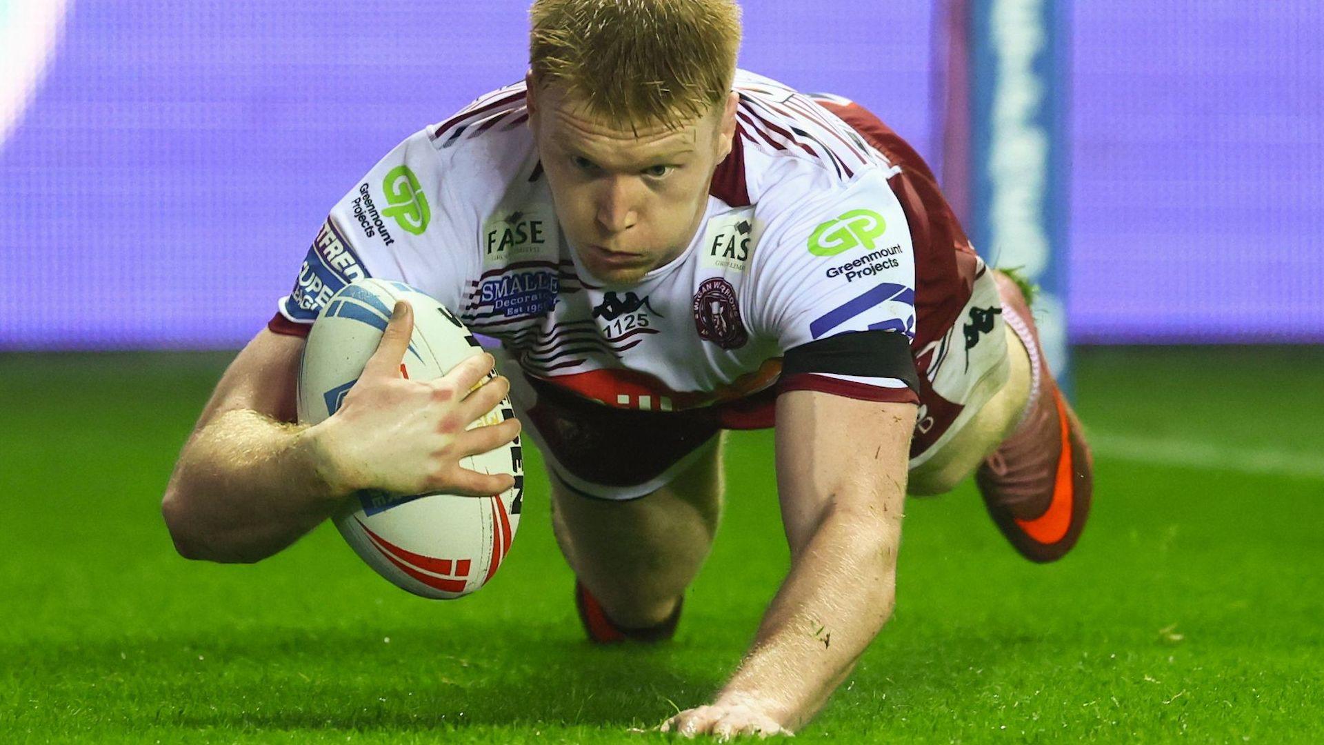 Zach Eckersley grips the ball in his right hand while diving to score for Wigan on a skiddy wet grass pitch.